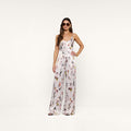 A woman in sunglasses stands confidently against a plain white background, wearing the Kimia Satin Pant—a white, sleeveless jumpsuit featuring a colorful butterfly print and straight-leg pants.