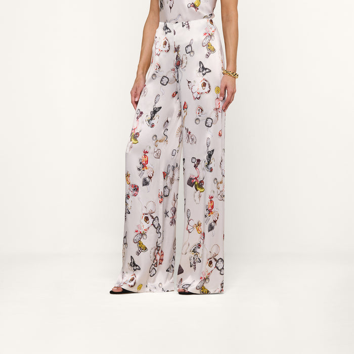 A person wearing the Kimia Satin Pant—a high-rise, silky white satin wide-leg pant featuring a colorful butterfly print—stands against a plain white background. Only the lower half of the body is visible.