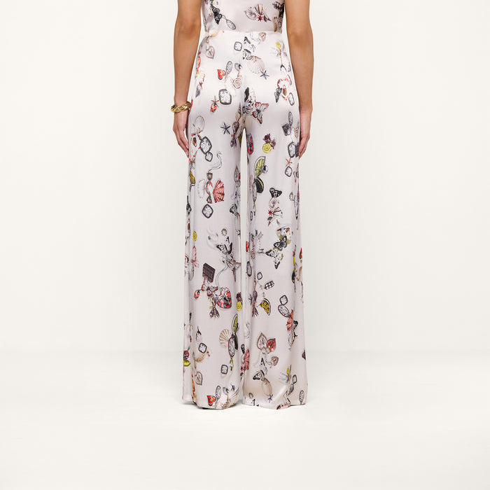 A woman is shown from the back wearing the Kimia Satin Pant—high-rise, wide-leg white pants featuring a colorful jewelry print—standing against a plain white background.