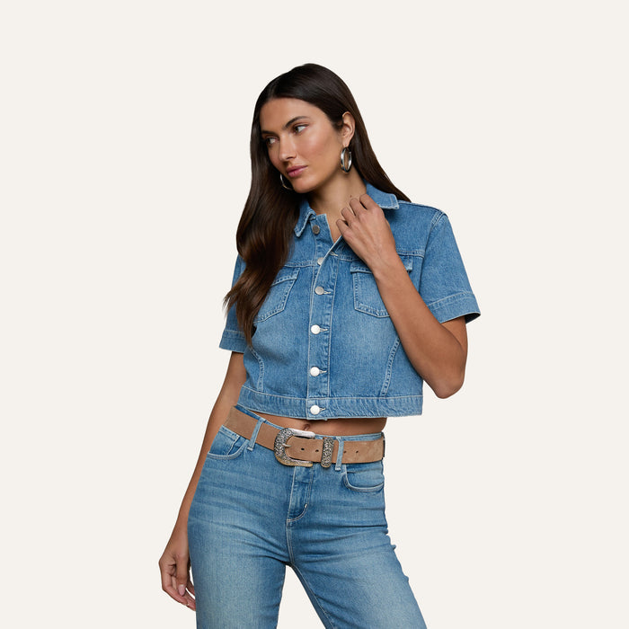 Wearing the Kirstie Jean Shirt Jacket, a woman with long dark hair pairs the short-sleeve cropped denim top with matching high-waisted jeans, a tan belt, and hoop earrings, standing against a plain light background.