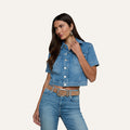 Wearing the Kirstie Jean Shirt Jacket, a woman with long dark hair pairs the short-sleeve cropped denim top with matching high-waisted jeans, a tan belt, and hoop earrings, standing against a plain light background.