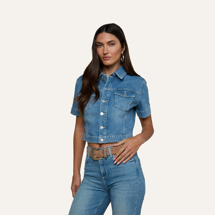 Wearing the Kirstie Jean Shirt Jacket—a cropped denim piece with short sleeves and front pockets—paired with high-waisted blue jeans, a woman stands against a plain light background.