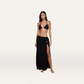 A woman stands barefoot against a plain, light background, wearing a black bikini top and the Klaire Cover-Up Skirt—a long, black wrap maxi skirt crafted from soft viscose chiffon. The skirt features an elegant tie closure at the waist for a customizable fit.