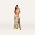 A woman stands barefoot with arms crossed against a plain white background, wearing the Klaire Cover-Up Skirt as part of a yellow floral two-piece outfit featuring a butterfly print and a wrap maxi skirt.