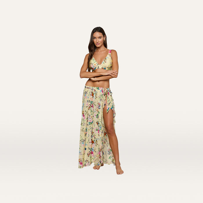 A woman stands barefoot with arms crossed against a plain white background, wearing the Klaire Cover-Up Skirt as part of a yellow floral two-piece outfit featuring a butterfly print and a wrap maxi skirt.