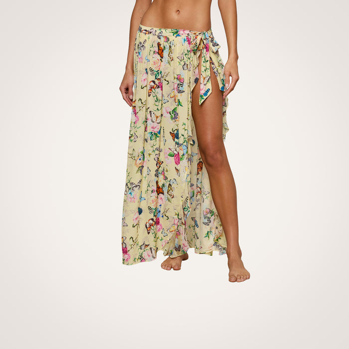 A woman stands barefoot against a plain white background, wearing the Klaire Cover-Up Skirt—a wrap maxi skirt in yellow with a delicate butterfly print and a high slit. Her upper body is not visible. The skirt is crafted from lightweight viscose chiffon, adding an elegant flow to the look.
