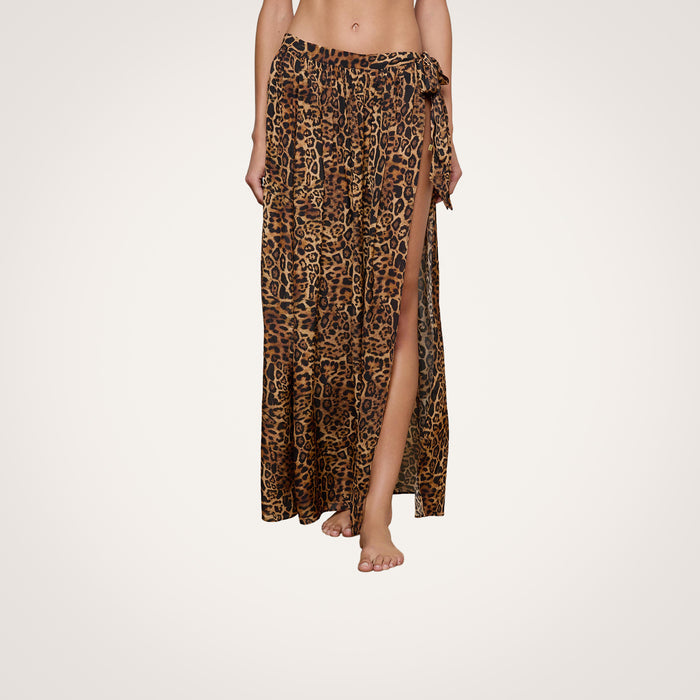 A person wearing the Klaire Cover-Up Skirt—a long, high-slit wrap skirt in leopard print—stands barefoot against a plain background. Made from airy viscose chiffon, the skirt features a relaxed fit and ties at the waist.