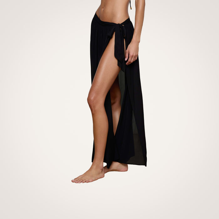 A woman wearing the Klaire Cover-Up Skirt, a black viscose chiffon wrap maxi skirt featuring a high slit and tie closure at the waist, stands barefoot against a plain light background.