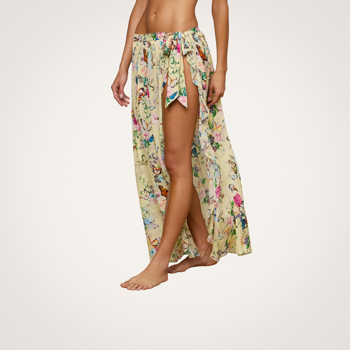 A person wearing the Klaire Cover-Up Skirt—a long, yellow wrap maxi skirt with a floral pattern, high slit, and crafted from airy viscose chiffon—stands barefoot against a plain white background. Only the lower body is visible.