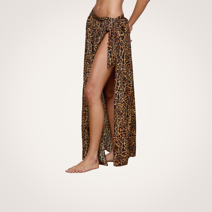 A person stands barefoot against a plain white background, wearing the Klaire Cover-Up Skirt—a high-slit wrap skirt in leopard print, crafted from airy viscose chiffon—with one leg and part of the midriff visible.