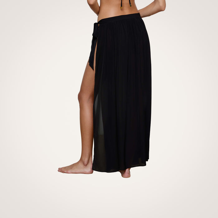 A person wearing the Klaire Cover-Up Skirt—a black sheer viscose chiffon wrap maxi skirt with a high side slit and tie closure—stands barefoot against a plain light background.