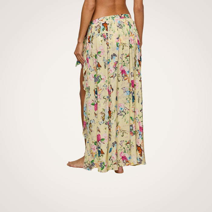 A person stands barefoot, wearing the Klaire Cover-Up Skirt—a long, light yellow viscose chiffon wrap maxi skirt featuring a high slit and delicate floral accents. The background is plain white, and only the lower half of their body is visible.