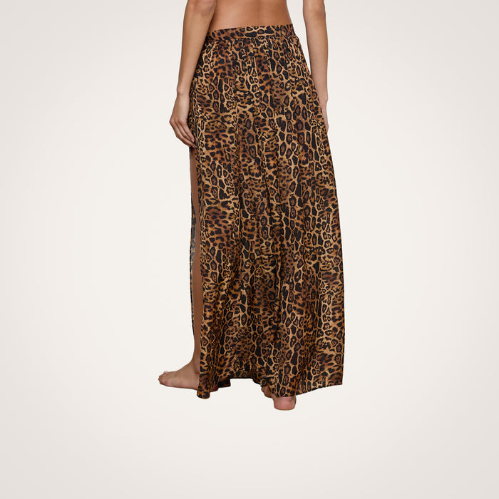 A woman stands barefoot, shown from the back against a plain light background, wearing the Klaire Cover-Up Skirt—a long, high-slit wrap skirt crafted from viscose chiffon and featuring a brown and black leopard print pattern.