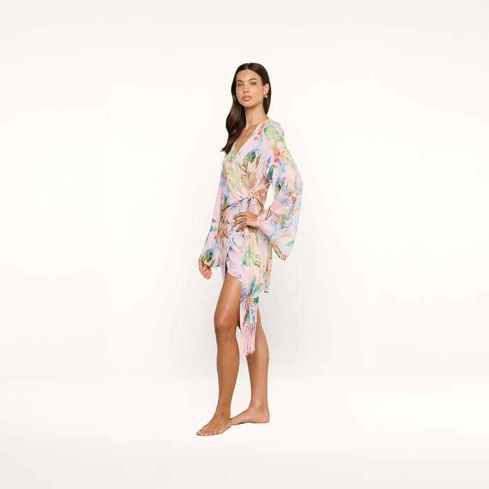 A woman stands barefoot in the Kohima Cover-Up Dress, a light pink floral kimono-style piece featuring long, loose bell sleeves and a wrap tie at the waist, evoking effortless resort style against a plain white background.