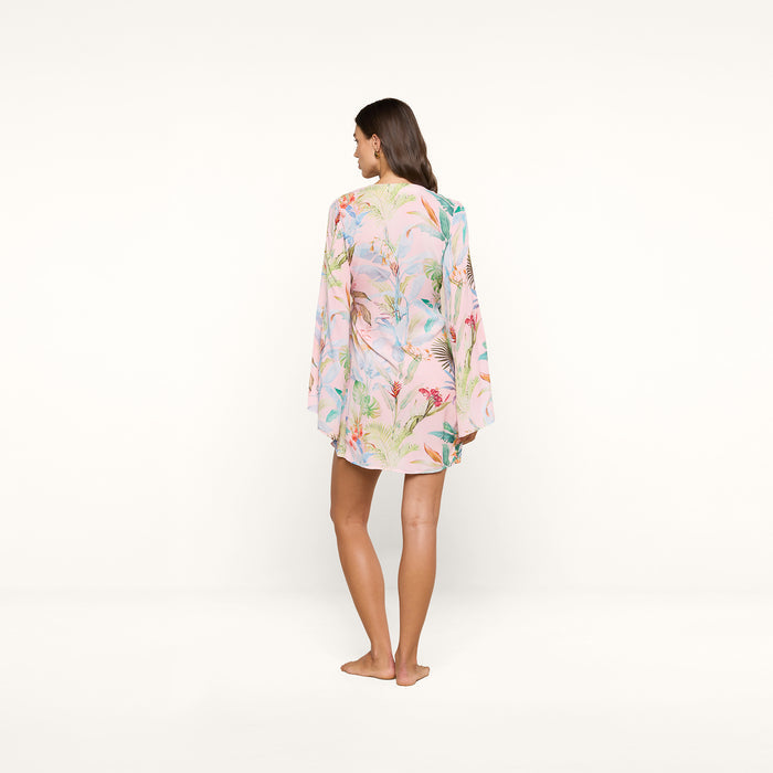 A woman stands barefoot facing away, wearing the Kohima Cover-Up Dress—a light, floral-patterned long-sleeve dress with subtle bell sleeves—set against a plain white background.