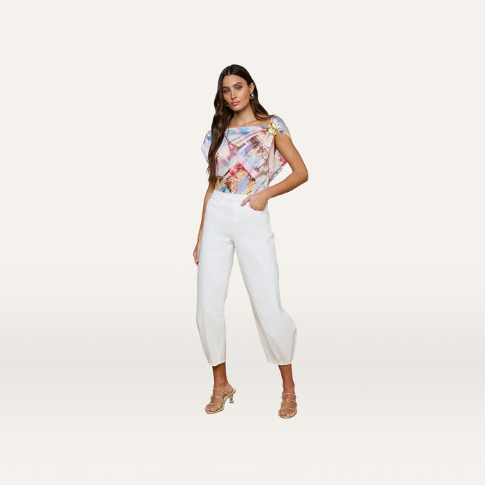 A woman in a colorful patterned short-sleeve top and Krista Tapered Jeans, paired with heeled sandals, stands against a plain light background with one hand in her pocket.