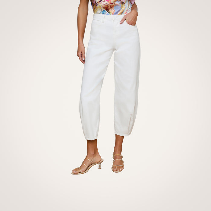 A woman models the Krista Tapered Jean, a white high-rise style with tapered legs, with her left hand in the pocket. She pairs it with strappy beige heels and a colorful patterned top against a plain light-colored background.