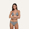 A woman wears the Kristin Triangle Bikini Top, a French Riviera swimwear-inspired black and white striped bikini featuring chain detail on the bottom, standing against a plain light background with her arms crossed.