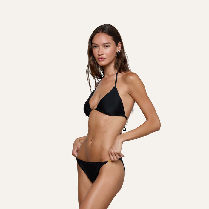 A woman models the Kristin Triangle Bikini Top, featuring adjustable ties and four-way stretch nylon, in black against a plain, light background. She stands slightly angled toward the camera, holding the side of her bikini bottom with one hand.