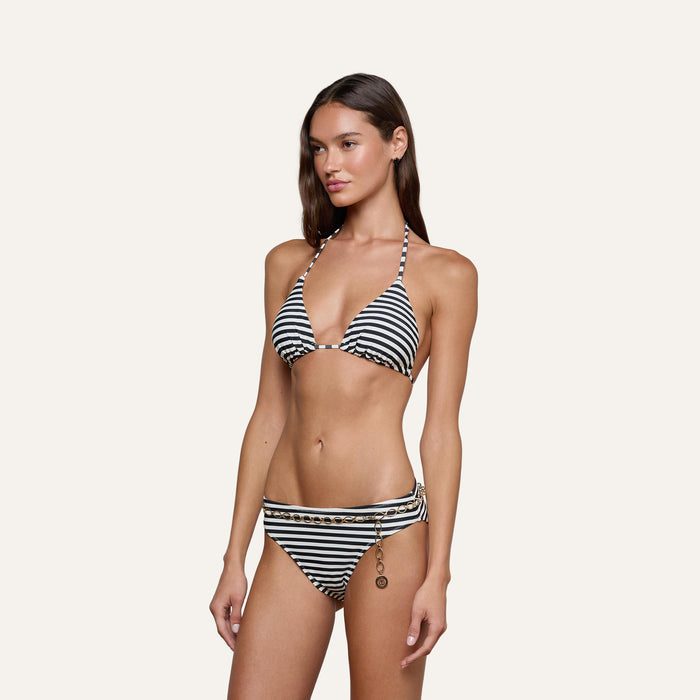 A woman wearing the Kristin Triangle Bikini Top, featuring black and white stripes and gold chain detail on the bottom, stands against a plain light background, embodying French Riviera swimwear style.