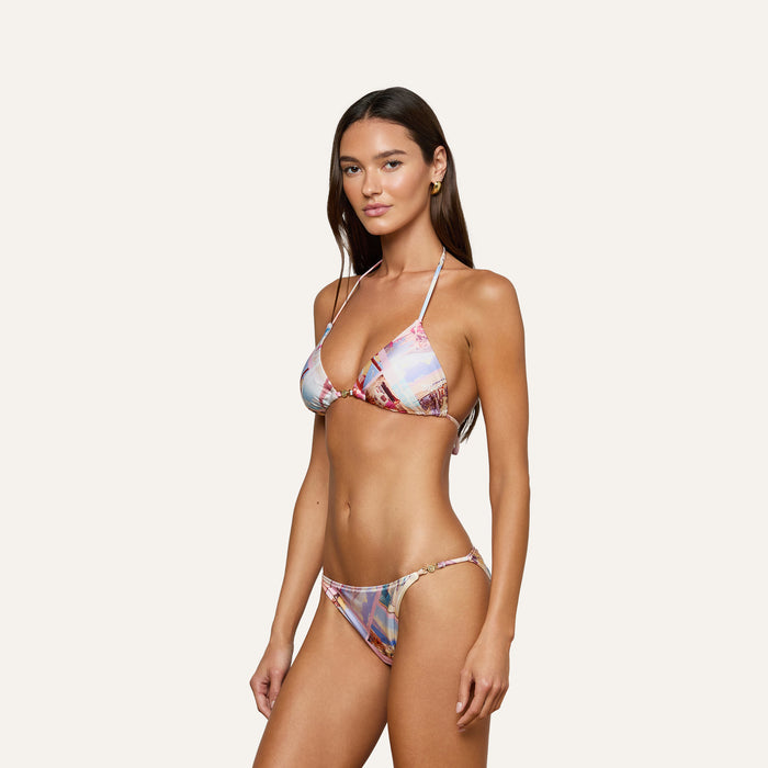 A woman with long brown hair models the Kristin Triangle Bikini Top, which features a vibrant postcard print cityscape. She stands at an angle against a plain light background. The bikini top offers adjustable coverage for a customizable fit.