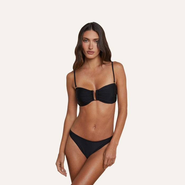 A woman stands against a plain, light background wearing the Krysta Bandeau Top in black with removable straps and matching bottoms.