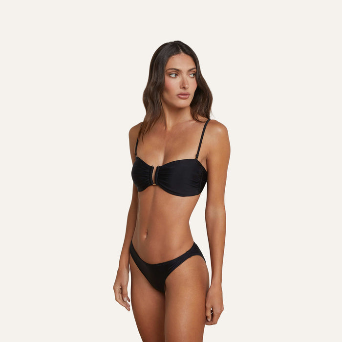 A woman wears the Krysta Bandeau Top, a black bikini top with thin, removable straps, as she looks to her left. The background is plain and light-colored.