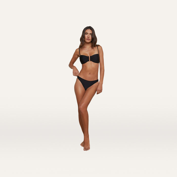 A woman stands barefoot against a plain white background, wearing the Krysta Bandeau Top—a black bikini with a bandeau-style silhouette featuring removable straps—paired with matching bottoms. She looks forward with a relaxed pose and neutral expression.