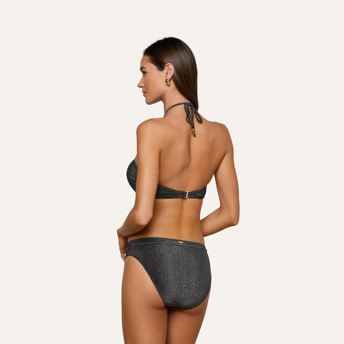 A woman stands sideways, facing away, wearing the Krysta Halter Bikini Top in metallic black with gold accents and hoop earrings. Her hands rest on her hips against a plain, light background.