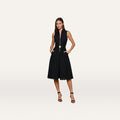 A woman stands against a plain white background wearing the Kylo Lace Shirt Dress—a sleeveless black knee-length dress with a collared neckline, gold belt, and black high-heeled sandals—perfect for an elegant desk-to-drinks look.