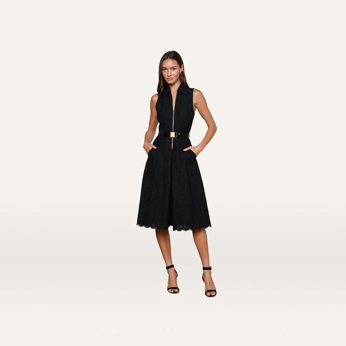 A woman stands against a plain white background wearing the Kylo Lace Shirt Dress—a sleeveless black knee-length dress with a collared neckline, gold belt, and black high-heeled sandals—perfect for an elegant desk-to-drinks look.