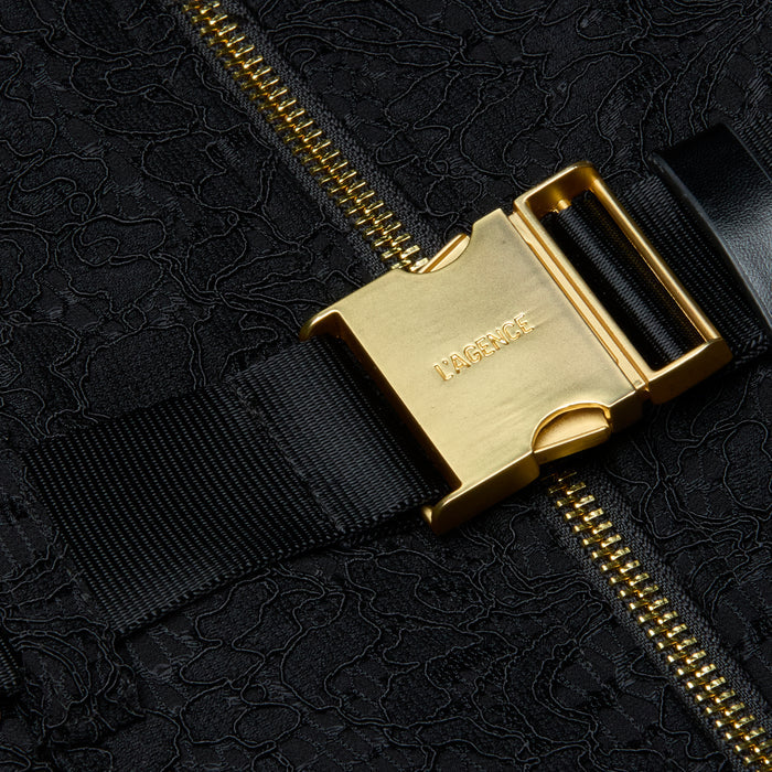 Close-up of a gold metal buckle with LAGENCE engraved, attached to a black strap and zippered, textured black fabric—an elegant detail that elevates the Kylo Lace Shirt Dress, perfect for adding polish to your favorite pocketed look or taking your style seamlessly from desk to drinks.