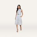 A woman stands against a plain white background, hands in pockets, wearing the Kylo Shirt Dress—a sleeveless blue lace print dress with a subtle floral pattern—paired with heeled sandals.