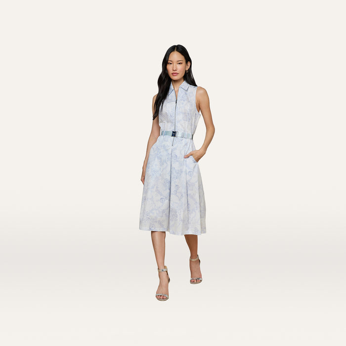 A woman stands against a plain white background, hands in pockets, wearing the Kylo Shirt Dress—a sleeveless blue lace print dress with a subtle floral pattern—paired with heeled sandals.