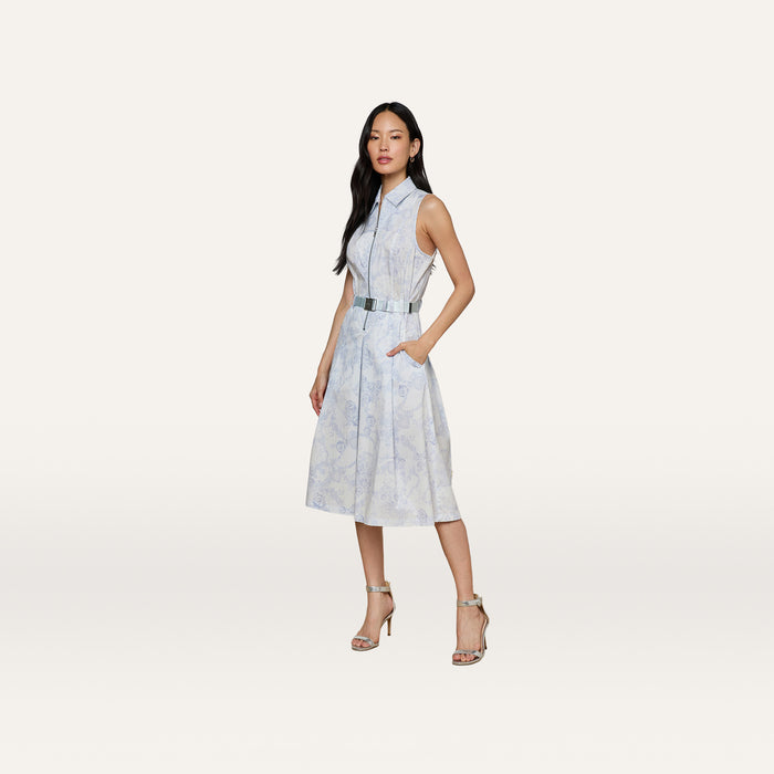 A woman stands against a plain white background wearing the Kylo Shirt Dress, a light blue cotton blend style featuring a collared neckline, belted waist, and practical pockets. She pairs the dress with high-heeled sandals and has long dark hair.