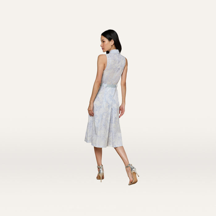 A woman models the Kylo Shirt Dress—a sleeveless, blue lace-print, knee-length dress—paired with high-heeled sandals. She stands against a plain light background, her back turned and glancing over her right shoulder.