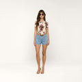 A woman wearing sunglasses, a floral print top, the Kyra Jean Short made from lightweight denim, and heeled sandals stands facing forward against a plain white background.