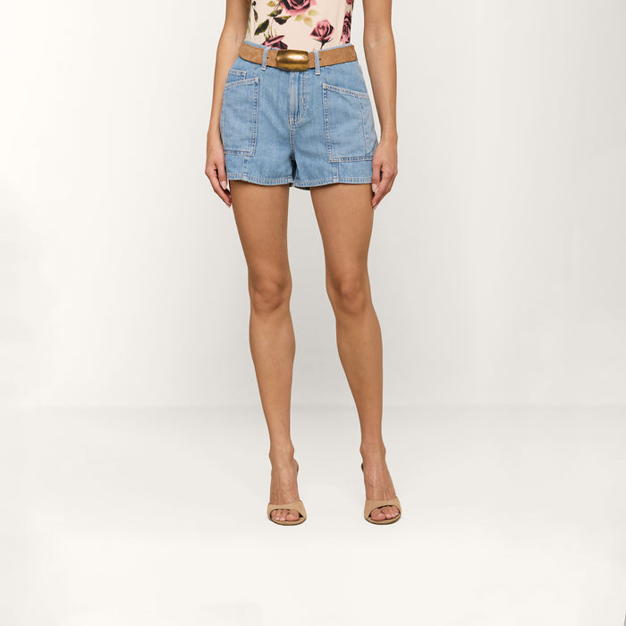 Wearing the Kyra Jean Short, a woman showcases high-rise, light blue jean shorts crafted from lightweight denim. She pairs them with a floral top, a beige belt, and beige sandals while standing against a plain white background.