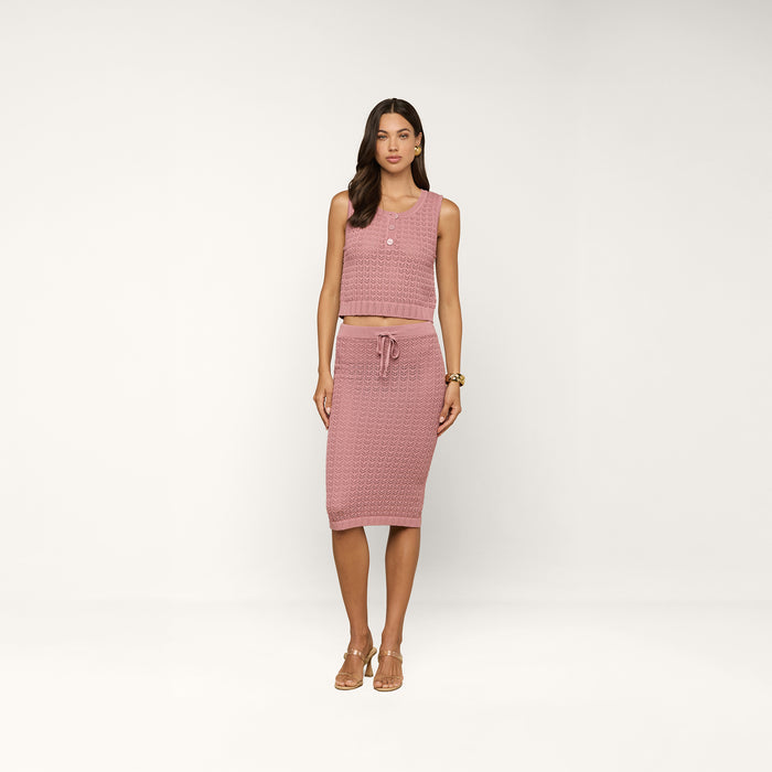 A woman stands in a studio against a plain white background, wearing the Lacen Pointelle Knit Skirt—a sleeveless, pink textured crop top paired with the matching midi-length skirt featuring a drawstring waist. She completes the look with tan sandals and gold jewelry.