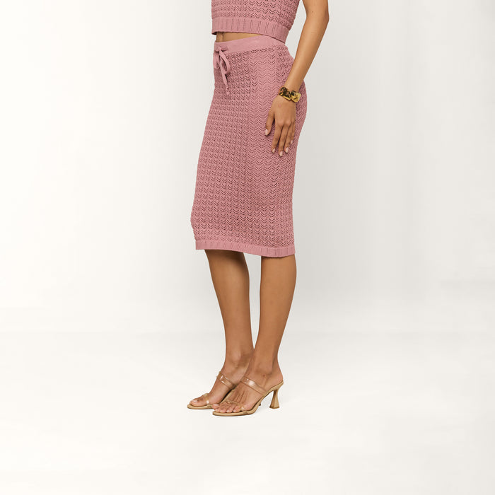 A woman stands sideways against a neutral background, wearing the Lacen Pointelle Knit Skirt—a pink, breathable midi-length knit skirt with a drawstring waist—paired with a matching top. She accessorizes with clear heeled sandals and a gold bracelet, her hands resting at her sides.