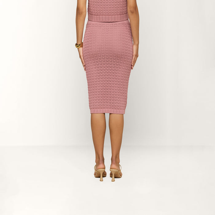 A woman stands facing away, wearing the Lacen Pointelle Knit Skirt in pink, styled as a knee-length, fitted, breathable knit piece. She pairs it with beige heeled sandals and a gold bracelet on her left wrist. The background is plain and light-colored.