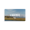 A white horse stands in a grassy field near water, set against a backdrop of mountains and blue sky. The image features the words “L’AGENCE Gift Card” in bold white text, evoking a sense of tranquility that mirrors the seamless experience of receiving thoughtful gift card redemption instructions.