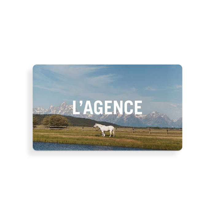 A white horse stands in a grassy field near water, set against a backdrop of mountains and blue sky. The image features the words “L’AGENCE Gift Card” in bold white text, evoking a sense of tranquility that mirrors the seamless experience of receiving thoughtful gift card redemption instructions.