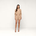 A woman with long brown hair and a neutral expression stands facing forward against a plain white background, wearing the Laina Eyelet Short—a lightweight, beige cotton eyelet short—paired with a matching floral-textured jacket and nude high heels.