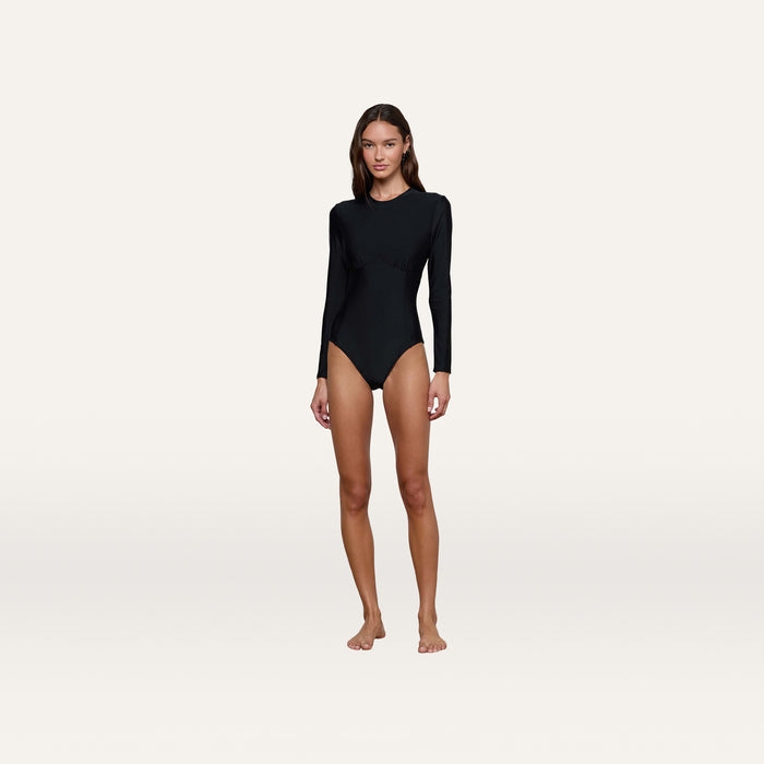 A woman stands barefoot on a plain background, wearing the Lambert Paddlesuit—a black long-sleeve paddlesuit crafted from four-way stretch nylon for a sculpted silhouette.