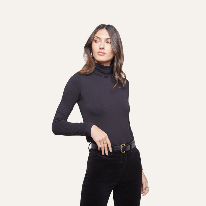 Wearing the Lani Turtleneck and black belted pants, a woman stands against a plain light background, looking to the side with one hand on her hip.