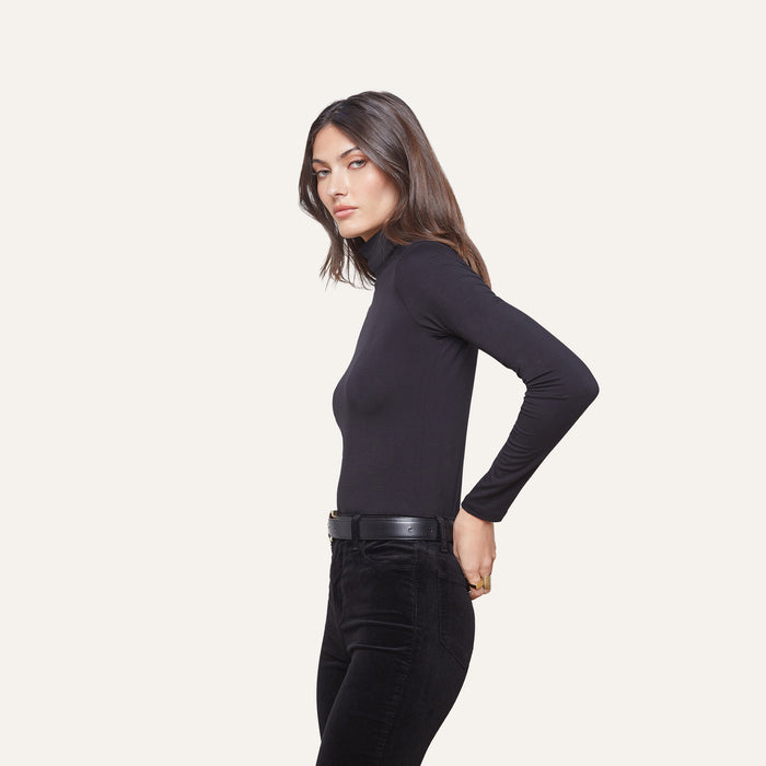 A woman stands in profile against a plain background, wearing the Lani Turtleneck in slim fit black, paired with black pants and a black belt, her hands behind her back and her dark hair down.