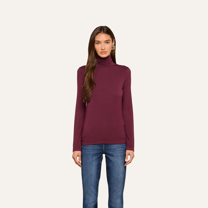 A woman with long brown hair wears the Lani Turtleneck in maroon and blue jeans, standing against a plain off-white background and looking straight at the camera with a neutral expression.