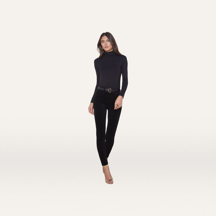 A woman stands against a plain white background, wearing the Lani Turtleneck in a slim fit, paired with black high-waisted pants with a belt and open-toe heels. She faces forward with a neutral expression and relaxed posture.