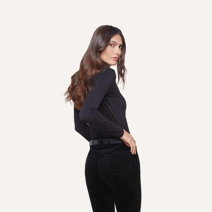 A woman with long brown hair stands sideways against a plain light background, wearing the Lani Turtleneck and black pants, looking over her shoulder.